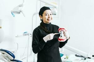 sorridente femmina dentista mostrando un' dentiera, dentista spiegando dente spazzolatura, femmina dentista mostrando un' dentiera e spazzola foto