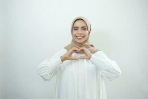 ritratto di sorridente asiatico musulmano donna mostrando cuore gesto isolato al di sopra di bianca sfondo foto