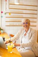 anziano donna utilizzando mobile Telefono mentre Lavorando su il computer portatile e potabile fresco arancia succo nel il bar foto