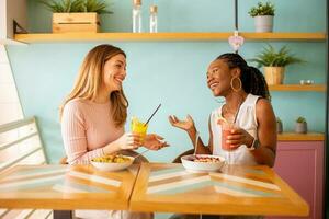 giovane nero e caucasico donna avendo bene volta, potabile fresco succhi di frutta e avendo salutare prima colazione nel il bar foto