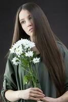 un' bellissimo giovane ragazza con naturale bellezza con lungo liscio capelli detiene un' mazzo di bianca crisantemi. foto