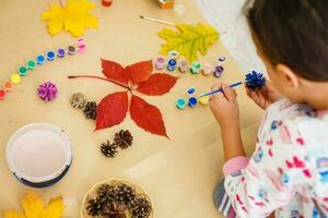 carino poco ragazza colorazione un' naturale cono abete per creare un' fiore, impostato di spazzole, vernici, naturale autunno materiali, creare divertimento e facile con figli, Fai da te per scuola materna foto