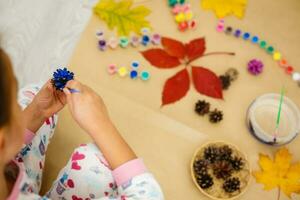 carino poco ragazza colorazione un' naturale cono abete per creare un' fiore, impostato di spazzole, vernici, naturale autunno materiali, creare divertimento e facile con figli, Fai da te per scuola materna foto