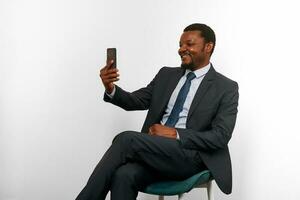 sorridente africano americano nero uomo nel attività commerciale completo da uomo seduta su sedia fabbricazione video chiamata attraverso Telefono foto