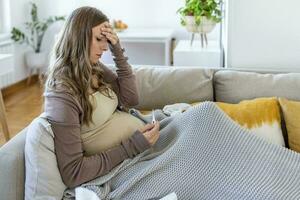 giovane incinta donna dire bugie nel letto, misurazione corpo temperatura, Tenere termometro e viso maschera foto