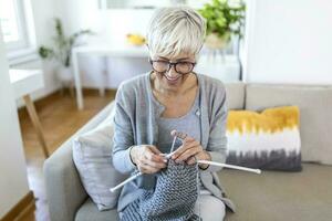 anziano donna nel bicchieri sedersi su divano a casa Sorridi Tenere maglieria aghi e filato maglia Abiti per amato quelli, preferito attività e passatempo, pensionato tranquillo spensierato vita concetto foto