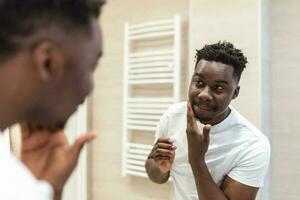 mattina igiene, bello uomo nel il bagno guardare nel specchio. riflessione di africano con barba guardare a specchio e toccante viso nel bagno governare foto