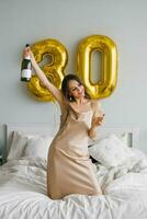 positivo donna celebra sua compleanno per trenta anni, seduta nel letto con un' bottiglia di Champagne e un' bicchiere nel sua mani foto