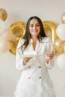 giovane caucasico donna Tenere un' torta con un' candela e un' bicchiere di Champagne nel onore di il trentesimo compleanno nel un' elegante bianca vestito con piume foto