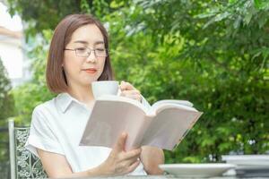 asiatico mezzo anziano donna nel bicchieri lettura un' libro nel Giardino dietro la casa foto