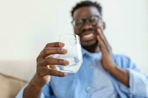 giovane uomo con sensibile denti e mano Tenere bicchiere di freddo acqua con Ghiaccio. assistenza sanitaria concetto. uomo potabile freddo bere, bicchiere pieno di ghiaccio cubi e si sente mal di denti, dolore foto