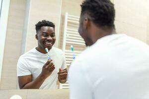 sorridente africano uomo con spazzolino pulizia denti e guardare specchio nel il bagno. uomo spazzolatura il suo denti nel mattina nel bagno. tipo nel pigiama spazzolatura denti a notte prima andando per dormire. foto