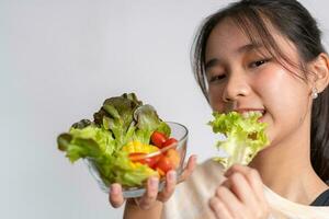 ritratto di un' contento giocoso asiatico ragazza mangiare fresco insalata a partire dal un' bicchiere ciotola dopo allenarsi a casa. giovane signora godendo salutare nutrizione e biologico cibo, avendo vegetariano pasto foto