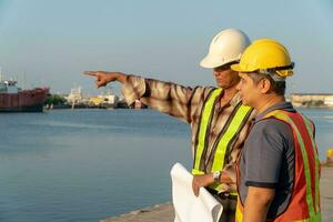 Due ingegneri si fermò indossare un' sicurezza casco. in piedi a il molo e Tenere il planimetria e consultare il Piano nel il costruzione. concetto di efficiente costruzione gestione. foto