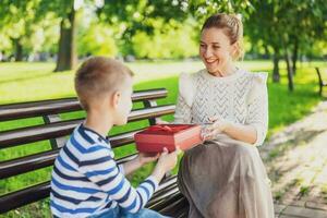 contento madre è seduta con sua figlio su panchina nel parco. ragazzo è dando un' presente per il suo madre. foto