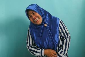 un' divertente di mezza età asiatico donna, nel un' blu hijab e a strisce camicia, scoppia in incontrollabile risata, isolato su un' blu sfondo foto
