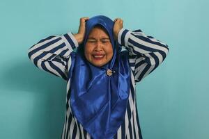 stressato asiatico donna nel blu hijab e a strisce camicia afferra testa, frustrato e dare le vertigini, contro blu sfondo foto