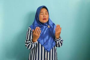 ritratto di un' di mezza età asiatico donna nel un' blu hijab e a strisce camicia è fabbricazione un' preghiere gesto con sua mani sollevato su. lei è isolato al di sopra di un' blu sfondo foto