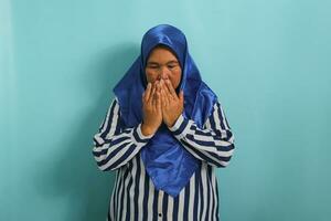 ritratto di un' di mezza età asiatico donna nel un' blu hijab e a strisce camicia è fabbricazione un' preghiere gesto con sua mani sollevato su. lei è isolato al di sopra di un' blu sfondo foto