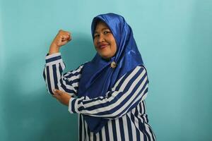 un eccitato di mezza età asiatico donna nel un' blu hijab e a strisce camicia Spettacoli un' forte gesto di sollevamento sua braccia e flessione sua muscoli, sorridente con orgoglio a il telecamera, isolato su blu sfondo. foto