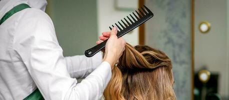 un' femmina parrucchiere è pettinatura il lungo Marrone capelli di un' giovane donna a un' salotto. foto