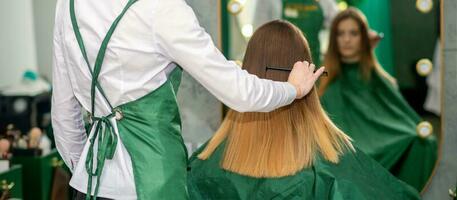 un' femmina parrucchiere è pettinatura il lungo Marrone capelli di un' giovane donna a un' salotto. foto