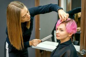 femmina parrucchiere messa in piega corto rosa capelli di il giovane bianca donna con mani e pettine nel un' capelli salone. foto