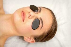 giovane donna ricevente facciale massaggio con nero minerale pietre su sua occhi nel un' spa. foto