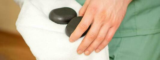 vicino su di del massaggiatore mano salviette nero massaggio pietre con un' bianca asciugamano. foto
