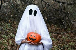 un' bambino vestito nel un' fantasma costume detiene un' Halloween zucca nel un' buio foresta. il bambino è avendo divertimento su Halloween. foto