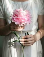 un' giovane donna nel un' bianca vestito teneramente detiene un' rosa peonia nel sua mani. foto