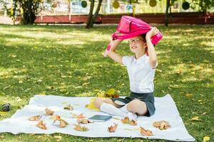 un' carino scolara con bicchieri sorrisi e detiene un' zaino sopra sua testa mentre seduta su un' coperta nel un' soleggiato autunno parco. indietro per scuola concetto foto