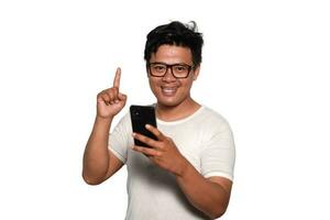 eccitato asiatico uomo con bicchieri indossare bianca maglietta sorridente mentre Tenere il suo Telefono, isolato di bianca sfondo foto