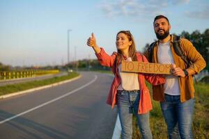 contento coppia autostop su ciglio della strada provando per fermare macchina. essi siamo Tenere cartone con iscrizione. foto