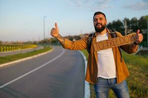 uomo è autostop su ciglio della strada provando per fermare macchina. lui è Tenere cartone con iscrizione. foto