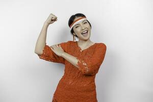 eccitato asiatico donna indossare un' rosso kebaya e fascia per capelli mostrando forte gesto di sollevamento sua braccia e muscoli sorridente con orgoglio. dell'Indonesia indipendenza giorno concetto. foto