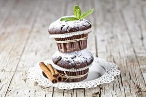 Muffin al cioccolato fondente con zucchero in polvere, bastoncini di cannella e foglia di menta su piastra bianca su tavola in legno rustico.
