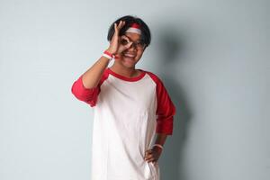 ritratto di attraente asiatico uomo nel maglietta con rosso e bianca nastro su testa, mostrando ok mano gesto e sorridente. isolato Immagine su grigio sfondo foto