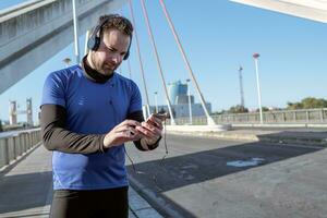 giovane uomo guardare a il suo cellula Telefono per ascolta per musica mentre in esecuzione attraverso un urbano la zona foto