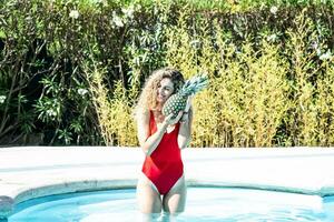 donna sorridente nel un' rosso costume da bagno dentro un' piscina abbracciare un' ananas foto