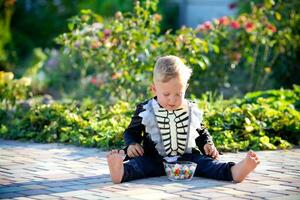 un' poco ragazzo si siede su il erba nel un' scheletro completo da uomo e mangia dolci. Halloween vacanza. copia spazio foto