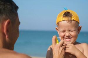 un' padre si applica protettivo crema per il suo figli maschi viso a il spiaggia. un' dell'uomo mano si applica protezione solare lozione su un' del bambino viso. foto