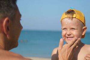 un' padre si applica protettivo crema per il suo figli maschi viso a il spiaggia. un' dell'uomo mano si applica protezione solare lozione su un' del bambino viso. foto