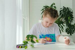 un' poco ragazza con un' gesso su sua braccio è fare sua compiti a casa. il bambino scrive compiti a casa nel un' taccuino. mano lesione. foto