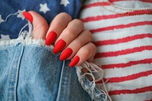 bellissimo femmina mani con un' luminosa rosso Opaco manicure su il sfondo di jeans. elegante ovale chiodo disegno, americano stile. estate manicure. copia spazio. foto
