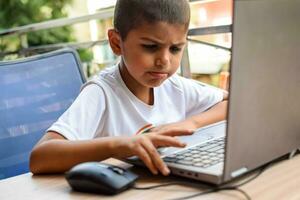 poco ragazzo seduta a tavolo utilizzando il computer portatile per in linea classe nel grado 1, bambino studiando su il computer portatile a partire dal casa per distanza apprendimento in linea formazione scolastica, scuola ragazzo bambini stile di vita concetto foto