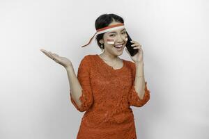 un' contento asiatico donna indossare rosso kebaya e fascia per capelli, Tenere sua Telefono, e puntamento copia spazio accanto suo, isolato di bianca sfondo. dell'Indonesia indipendenza giorno foto