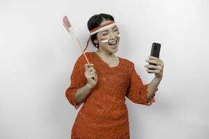 un' contento asiatico donna indossare rosso kebaya e fascia per capelli, Tenere sua Telefono e dell'Indonesia bandiera, isolato di bianca sfondo. dell'Indonesia indipendenza giorno foto