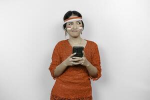 un' inteligente giovane donna indossare fascia per capelli e rosso kebaya è guardare a parte per un idea su copia spazio mentre Tenere sua Telefono, isolato di bianca sfondo. dell'Indonesia indipendenza giorno. foto
