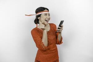 un' contento asiatico donna indossare rosso kebaya e fascia per capelli, Tenere sua Telefono, e Esprimere a gesti amore cartello di sua dita mostrando tenero sensazione, isolato di bianca sfondo. dell'Indonesia indipendenza giorno foto